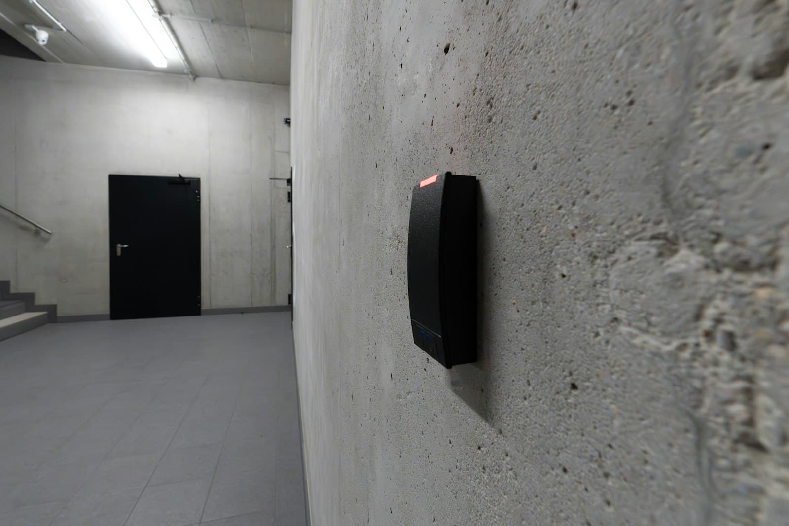 A black access reader on a concrete wall, a dark door beyond it.